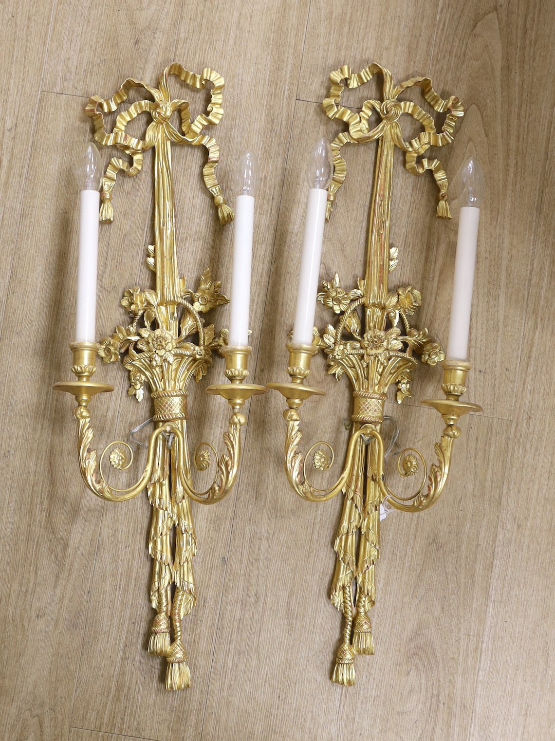 A pair of Louis XVI style carved giltwood wall lights, with ribbon crests and flowers in basket stems, over tasselled bases, height 85cm